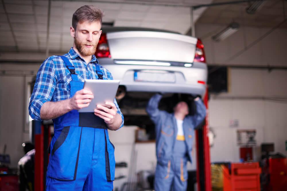 Ein Mechaniker nutzt ein Tablet für die schrittweise Schadensabwicklung, während ein Kollege ein Auto auf der Hebebühne überprüft