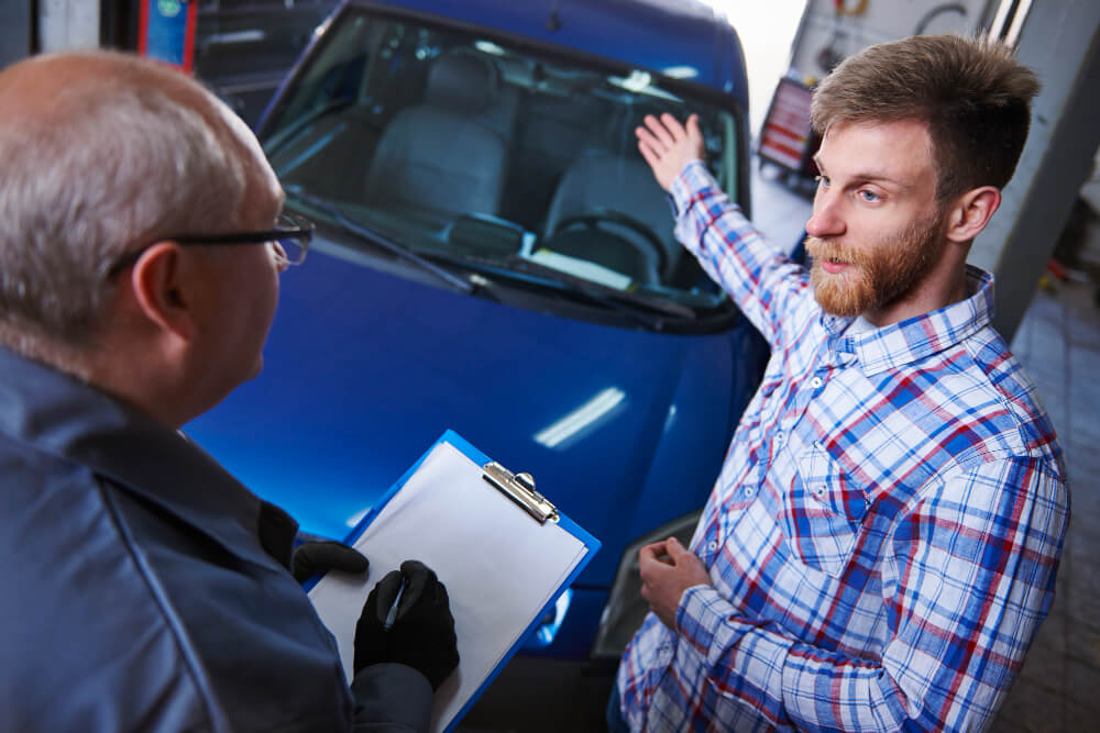 Ein Mann in einem karierten Hemd zeigt auf ein blaues Auto und bespricht den Kostenvoranschlag mit einem Mechaniker, der ein Klemmbrett in der Hand hält.