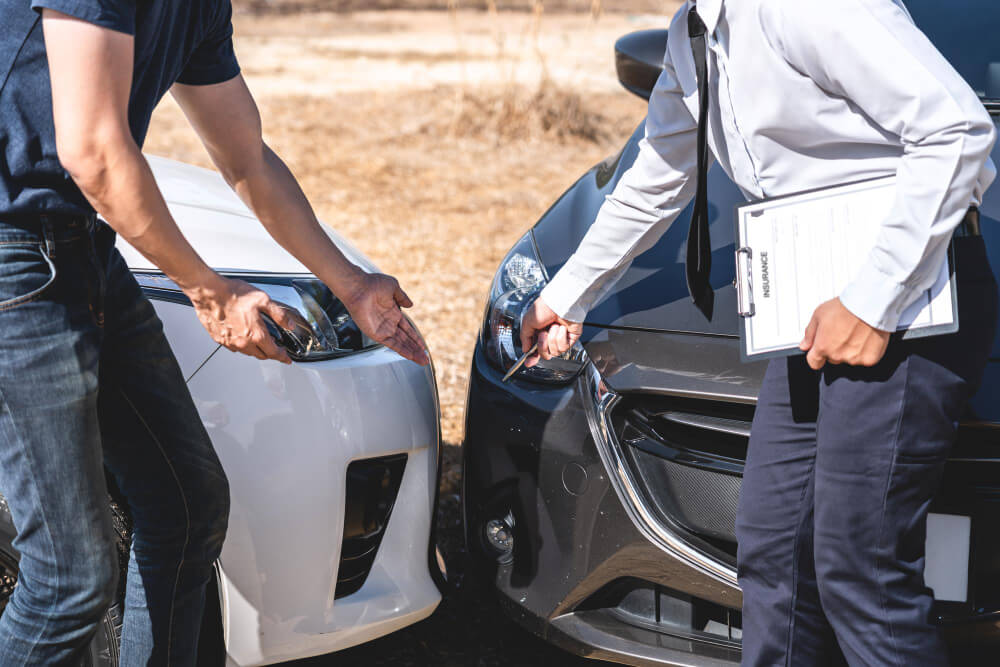 Zwei Personen begutachten einen Unfallschaden am Auto; eine hält ein Klemmbrett bereit, um einen Kfz-Sachverständigen zu beauftragen