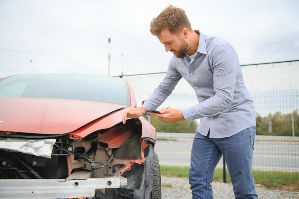 Ein Mann fotografiert den Schaden an einem roten Auto im Freien, wahrscheinlich für ein Gutachten oder die gegnerische Versicherung